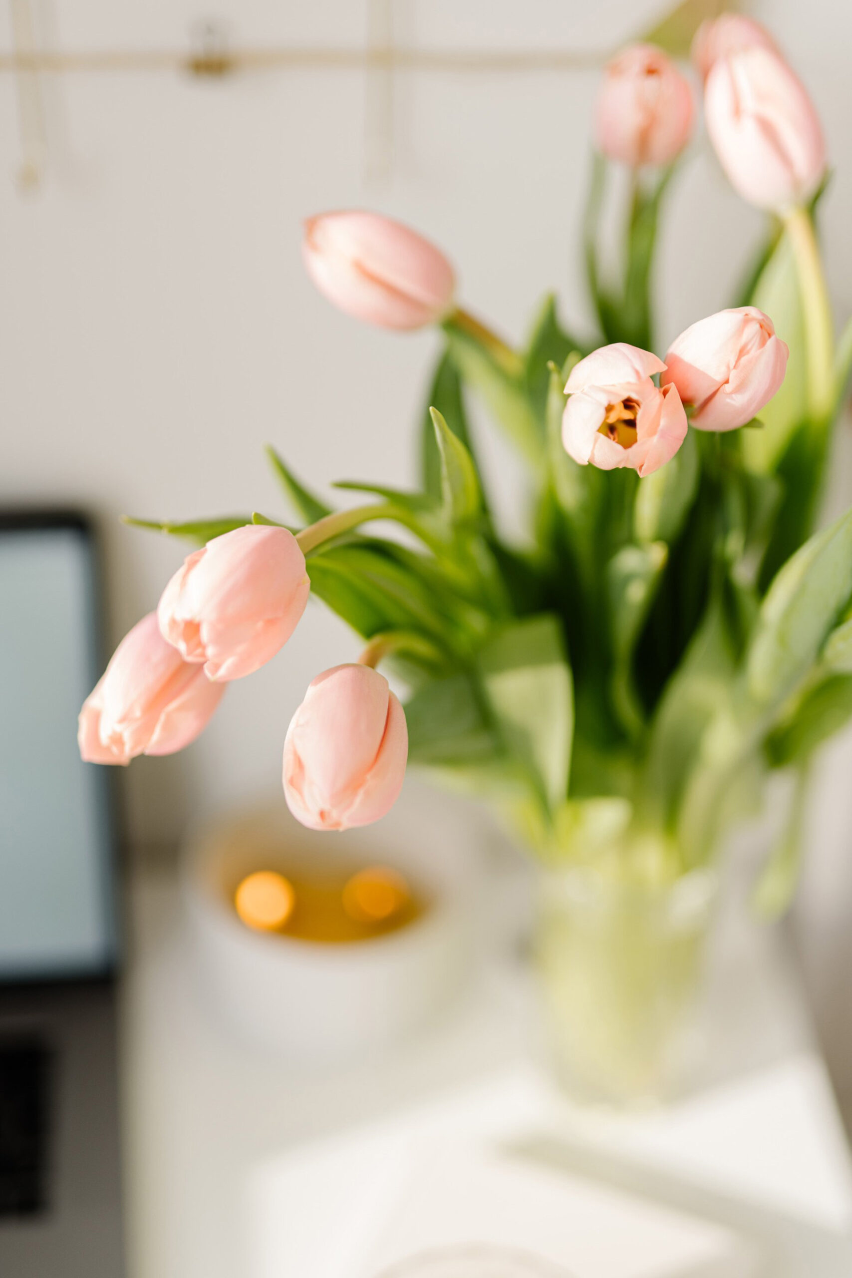 A bouquet of delicate pink tulips in a clear vase, set against a softly blurred background with a laptop and a candle, evoking a serene workspace atmosphere.